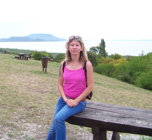 Mosolygó nő ül egy fából készült padon, háttérben a Balaton.